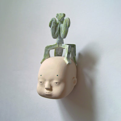 Whittled wooden sculpture of a floating head with a blank expression. Atop its head is a large green mantis, standing up.