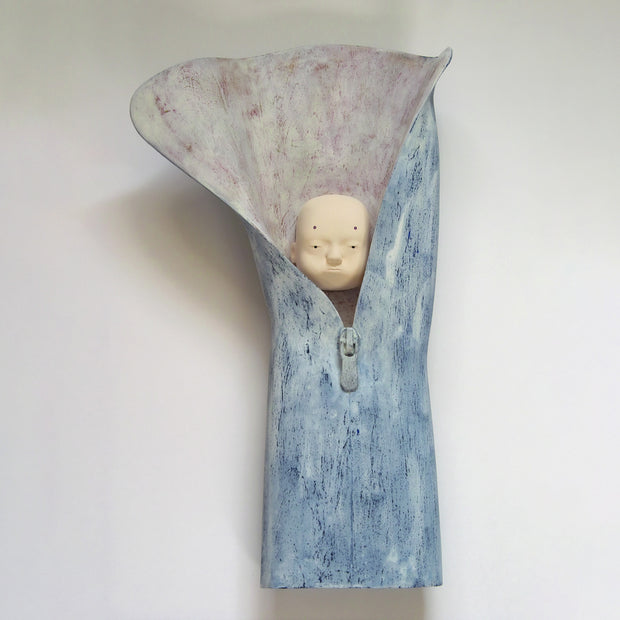 Carved wooden sculpture of a floating bald head with a blank expression. It is inside of a partially unzipped sleeve of blue material.