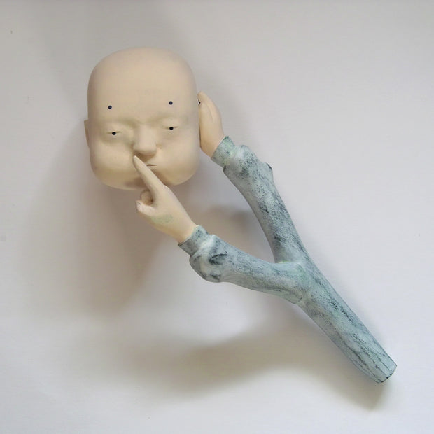 Carved wooden sculpture of a floating bald head with a blank expression. Attached to it is a stick like item with 2 sleeved arms coming out of it. One hand covers the head's ear and the other puts a finger up to its mouth, shushing it.