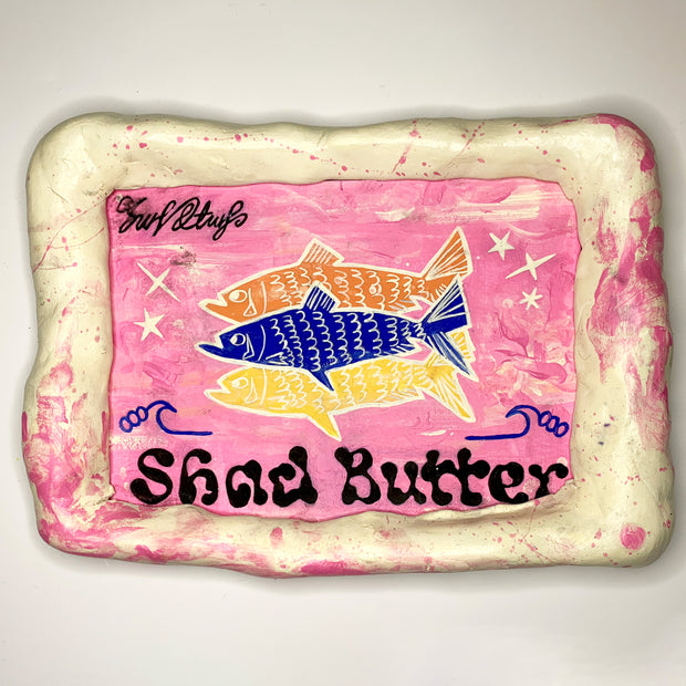 Painting encased in cream and pink colored clay frame, of 3 solid colored illustrations of fish on a light pink background. "Shad Butter" is written stylistically in black along the bottom.