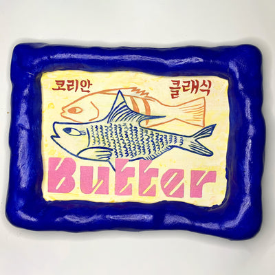 Painting encased in deep blue clay frame, of 2 line art illustrations of fish on a light yellow background. "Butter" is written stylistically in pink along the bottom and small Korean writing is up top.