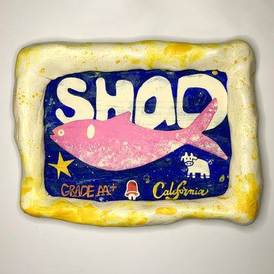 Painting encased in a cream colored and yellow clay frame, of a pink fish on dark blue background. "Shad" is written stylistically in white behind the fish with "Grade AA+ and "California" along the bottom with illustrations of a cow and a bell.