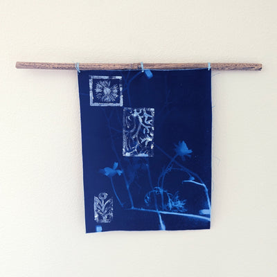 Wall hanging on a piece of driftwood of a blue cyanotype on fabric. Flowers and seaweed are imprinted.