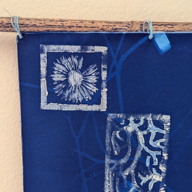 Wall hanging on a piece of driftwood of a blue cyanotype on fabric. Flowers and seaweed are imprinted.