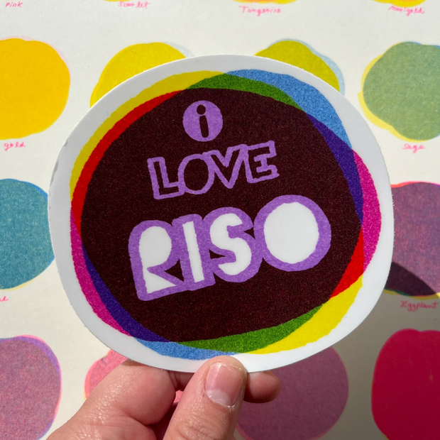 Rounded sticker that reads "I Love Riso" in purple and white font, on a dark background made up of several colors overlayed atop one another.