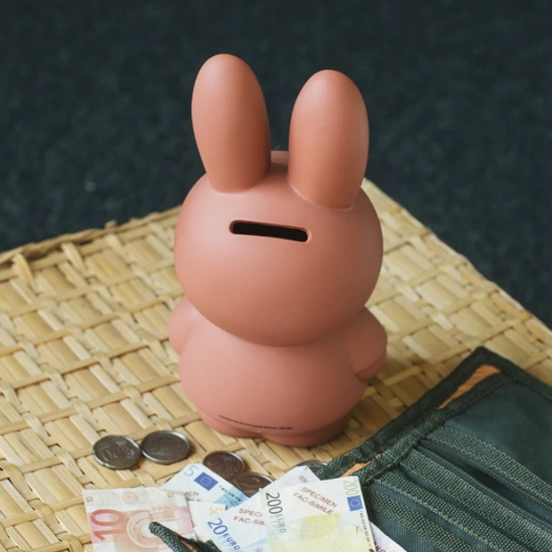 Miffy shaped coin bank, completely colored a terracotta orangish-brown with gray eyes and mouth. Seen from the back with a large coin slot on the back of her head.