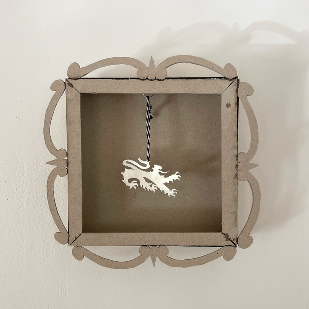 Silver pennant of a cat that resembles a dragon, akin to the one on the Welsh flag. The pennant hangs from a black and white striped twine, within a hand build cardboard shadow box.