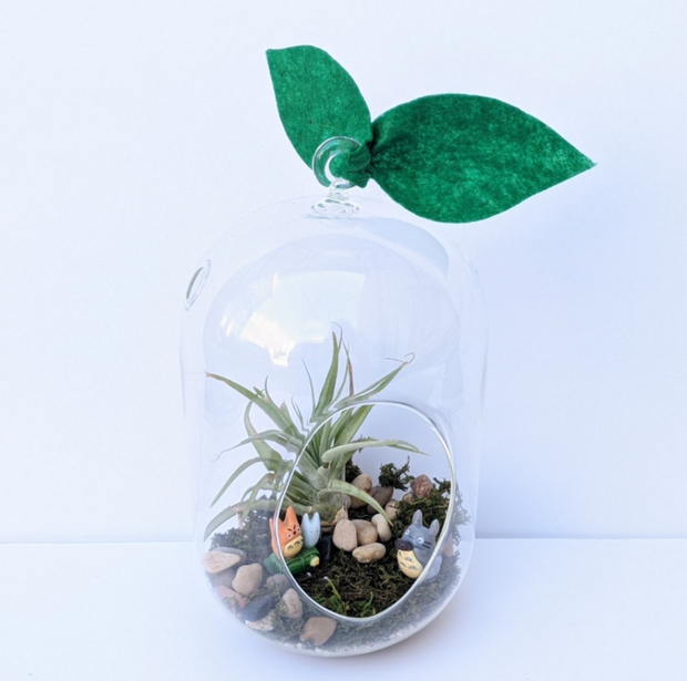 A tall glass terrarium has two green, felt leaves attached to the hanging ring at the top. There is a spikey, air plant inside along side some river pebbles, green moss carpet and three totoro figurines. One is frowning. Another is holding an ocarina