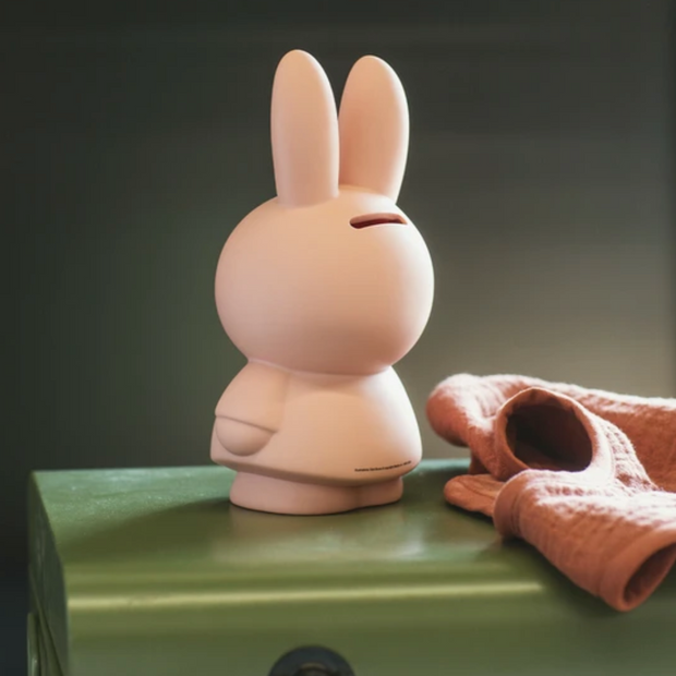 Miffy shaped coin bank, completely colored a smoky pink with gray eyes and mouth. Seen from the back to show the coin slot on the top of her head.