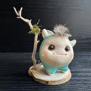 Ceramic sculpture of a stylized cute troll monster, with a large face and rounded bluebody with feet and no arms. It has a fluff atop its head and stands on a wooden base with a branch behind it and crystals.