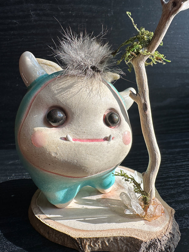 Ceramic sculpture of a stylized cute troll monster, with a large face and rounded bluebody with feet and no arms. It has a fluff atop its head and stands on a wooden base with a branch behind it and crystals.