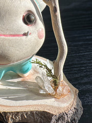 Ceramic sculpture of a stylized cute troll monster, with a large face and rounded bluebody with feet and no arms. It has a fluff atop its head and stands on a wooden base with a branch behind it and crystals.