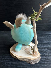 Ceramic sculpture of a stylized cute troll monster, with a large face and rounded bluebody with feet and no arms. It has a fluff atop its head and stands on a wooden base with a branch behind it and crystals.