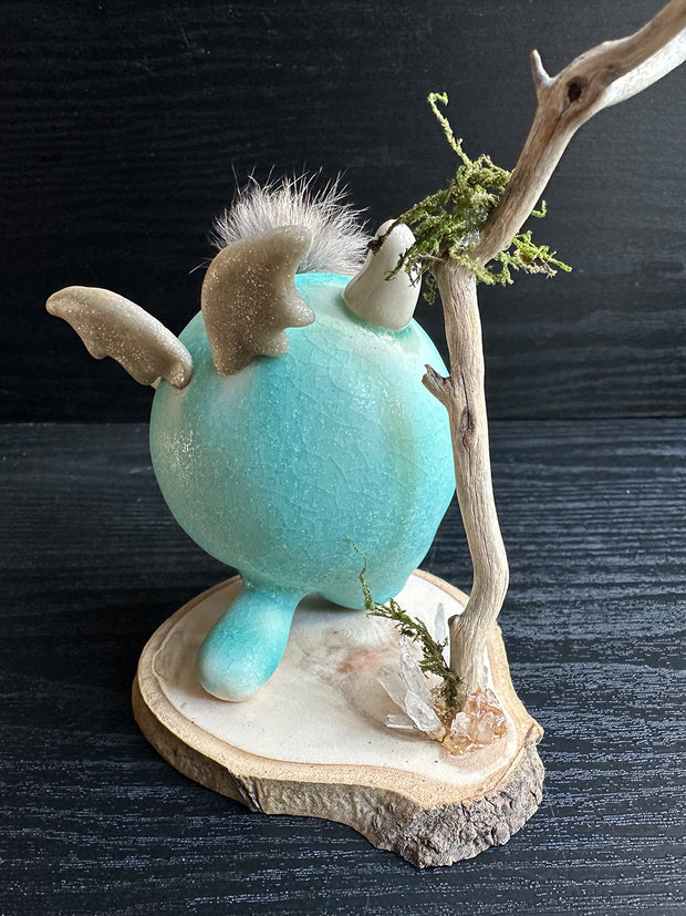 Ceramic sculpture of a stylized cute troll monster, with a large face and rounded bluebody with feet and no arms. It has a fluff atop its head and stands on a wooden base with a branch behind it and crystals.