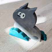 Wooden die cut sculpture of a gray creature with short pointed ears and large cartoon eyes and a small black nose. It looks over its shoulder with an inquisitive look and is between 2 small teal bushes.