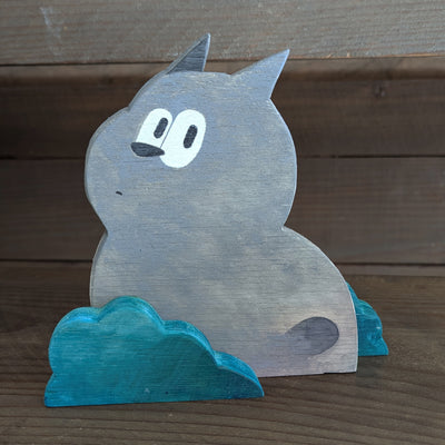 Wooden die cut sculpture of a gray creature with short pointed ears and large cartoon eyes and a small black nose. It looks over its shoulder with an inquisitive look and is between 2 small teal bushes.