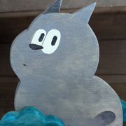 Wooden die cut sculpture of a gray creature with short pointed ears and large cartoon eyes and a small black nose. It looks over its shoulder with an inquisitive look and is between 2 small teal bushes.