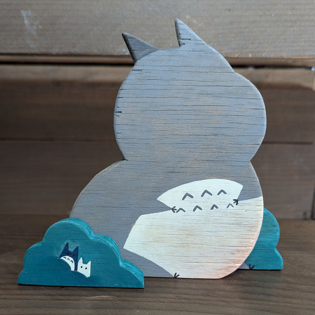 Wooden die cut sculpture of a gray creature with short pointed ears and large cartoon eyes and a small black nose. It looks over its shoulder with an inquisitive look and is between 2 small teal bushes.