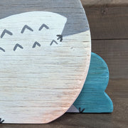 Wooden die cut sculpture of a gray creature with short pointed ears and large cartoon eyes and a small black nose. It looks over its shoulder with an inquisitive look and is between 2 small teal bushes.