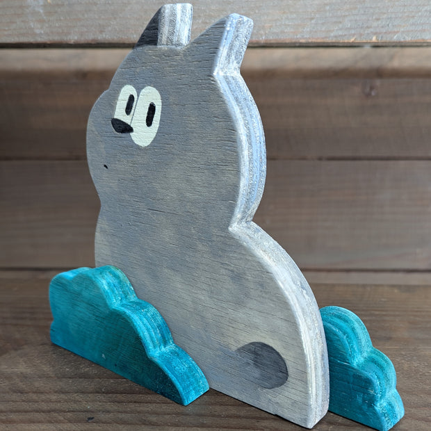 Wooden die cut sculpture of a gray creature with short pointed ears and large cartoon eyes and a small black nose. It looks over its shoulder with an inquisitive look and is between 2 small teal bushes.