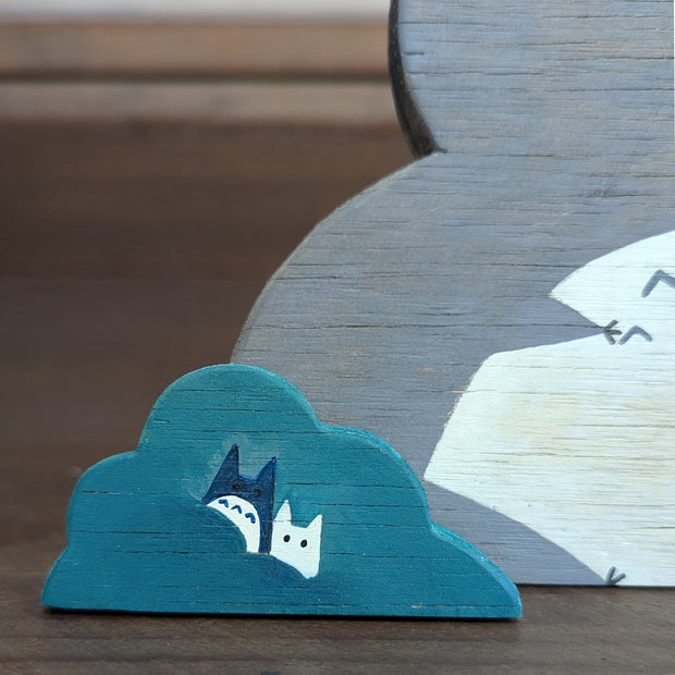 Wooden die cut sculpture of a gray creature with short pointed ears and large cartoon eyes and a small black nose. It looks over its shoulder with an inquisitive look and is between 2 small teal bushes.