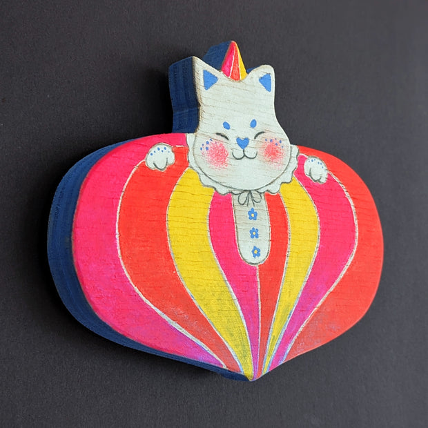 Painted wooden die cut sculpture of a white cat with a closed eye smile, dressed in a very puffy circus costume - pink, red and yellow striping that tapers in at the bottom like a blown up balloon. 