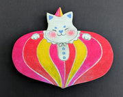 Painted wooden die cut sculpture of a white cat with a closed eye smile, dressed in a very puffy circus costume - pink, red and yellow striping that tapers in at the bottom like a blown up balloon. 