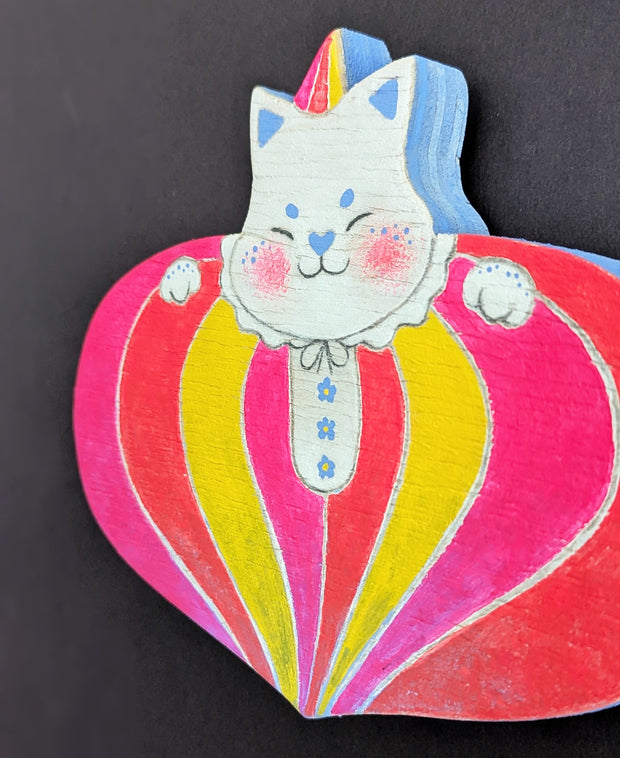 Painted wooden die cut sculpture of a white cat with a closed eye smile, dressed in a very puffy circus costume - pink, red and yellow striping that tapers in at the bottom like a blown up balloon. 