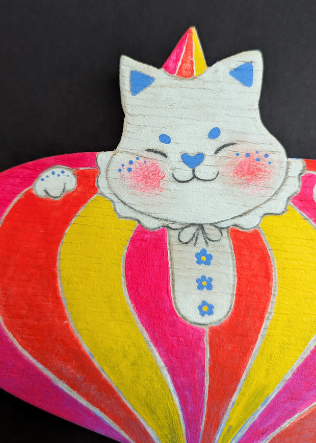 Painted wooden die cut sculpture of a white cat with a closed eye smile, dressed in a very puffy circus costume - pink, red and yellow striping that tapers in at the bottom like a blown up balloon. 