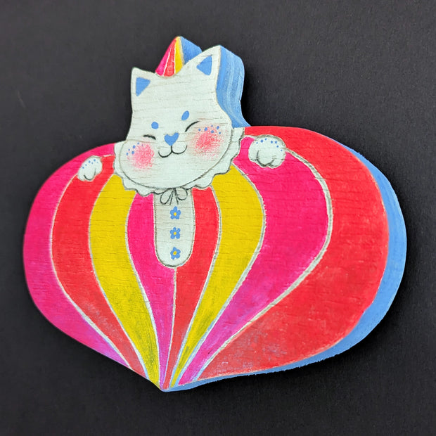 Painted wooden die cut sculpture of a white cat with a closed eye smile, dressed in a very puffy circus costume - pink, red and yellow striping that tapers in at the bottom like a blown up balloon. 