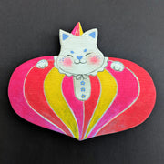 Painted wooden die cut sculpture of a white cat with a closed eye smile, dressed in a very puffy circus costume - pink, red and yellow striping that tapers in at the bottom like a blown up balloon. 