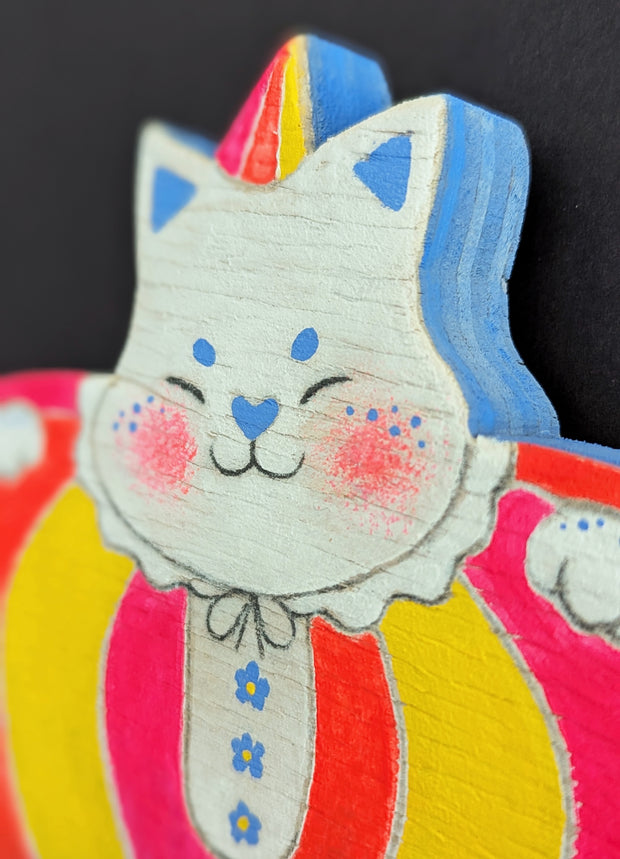 Painted wooden die cut sculpture of a white cat with a closed eye smile, dressed in a very puffy circus costume - pink, red and yellow striping that tapers in at the bottom like a blown up balloon. 