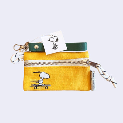 Small yellow canvas pouch with a zip closure, featuring a small illustration in the left corner of riding on a skateboard, going fast with speed lines behind him. It is attached to a green fabric lanyard.