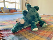 Fluffy plush doll of a sage green dinosaur or crocodile with large plastic cartoon eyes. It has teeth peeking out of its mouth and a long tail.