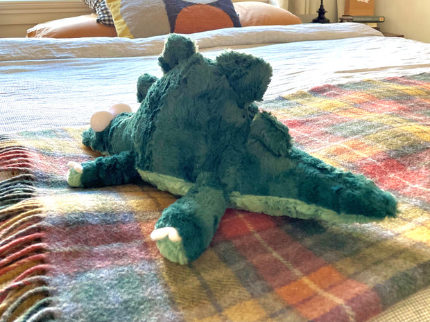 Fluffy plush doll of a sage green dinosaur or crocodile with large plastic cartoon eyes. It has teeth peeking out of its mouth and a long tail.