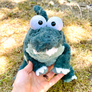 Fluffy plush doll of a sage green dinosaur or crocodile with large plastic cartoon eyes. It has teeth peeking out of its mouth and a long tail.