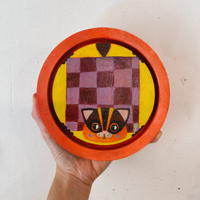 Painting on rounded inlayed wood panel of a flying squirrel, with a purple checkerboard pattern and an orange face. Background is yellow and border is orange.