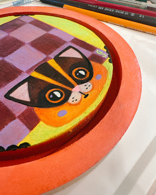 Painting on rounded inlayed wood panel of a flying squirrel, with a purple checkerboard pattern and an orange face. Background is yellow and border is orange.