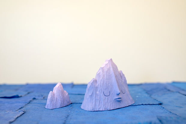 Pair of 2 carved wooden sculptures, each of small, purple mountain ranges. One has a closed eye, calm expression with subtle white stars around it. The other is faceless and smaller in size.