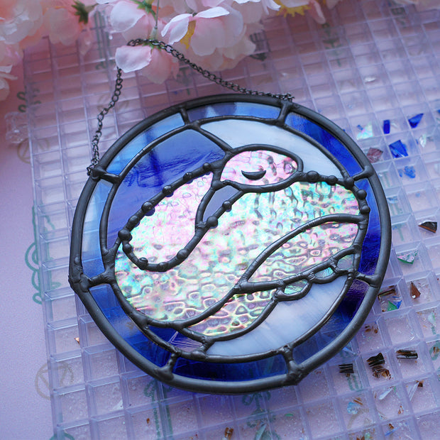 Stained glass circular hanging artwork of an iridescent snake, curled into itself and resting its head atop its body with closed eyes. Background is white and blue.