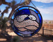 Stained glass circular hanging artwork of an iridescent snake, curled into itself and resting its head atop its body with closed eyes. Background is white and blue.