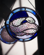 Stained glass circular hanging artwork of an iridescent snake, curled into itself and resting its head atop its body with closed eyes. Background is white and blue.