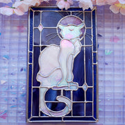 Stained glass rectangle shape hanging of a white cat sitting on a sill, only the silhouette visible aside from a purplish moon behind it and its many silver whiskers. Behind it are blue stained glass pieces.