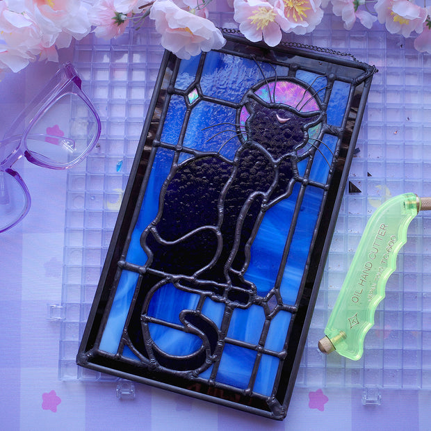 Stained glass rectangle shape hanging of a black cat sitting on a sill, only the silhouette visible aside from a crescent moon on its forehead. Behind it are blue stained glass pieces and a dark purple border.