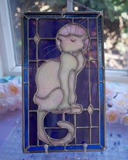 Stained glass rectangle shape hanging of a white cat sitting on a sill, only the silhouette visible aside from a purplish moon behind it and its many silver whiskers. Behind it are blue stained glass pieces.