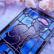 Stained glass rectangle shape hanging of a black cat sitting on a sill, only the silhouette visible aside from a crescent moon on its forehead. Behind it are blue stained glass pieces and a dark purple border.