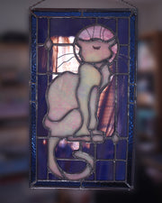 Stained glass rectangle shape hanging of a white cat sitting on a sill, only the silhouette visible aside from a purplish moon behind it and its many silver whiskers. Behind it are blue stained glass pieces.