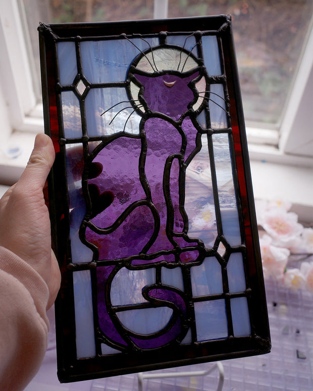 Stained glass rectangle shape hanging of a black cat sitting on a sill, only the silhouette visible aside from a crescent moon on its forehead. Behind it are blue stained glass pieces and a dark purple border.