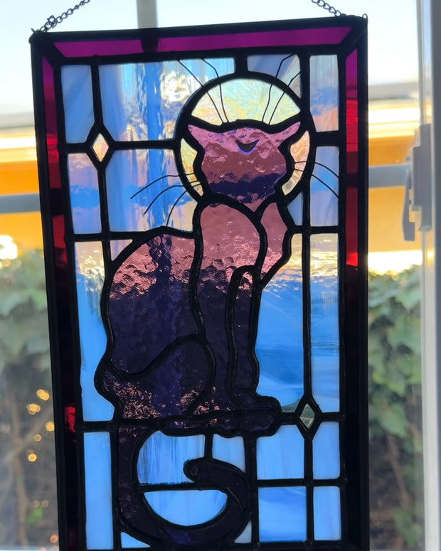 Stained glass rectangle shape hanging of a black cat sitting on a sill, only the silhouette visible aside from a crescent moon on its forehead. Behind it are blue stained glass pieces and a dark purple border.