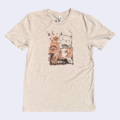 Tan t-shirt with an illustration of a cat reading a book with flowers and a small bird holding a flower in its beak.